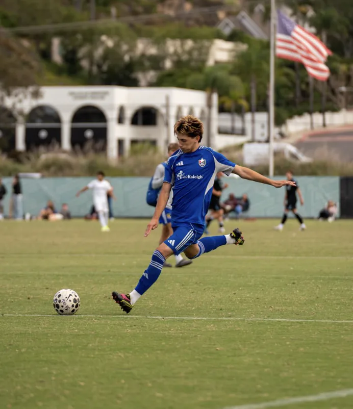 Soccer player in a blue kit strikes the ball with an American flag beyond the field.